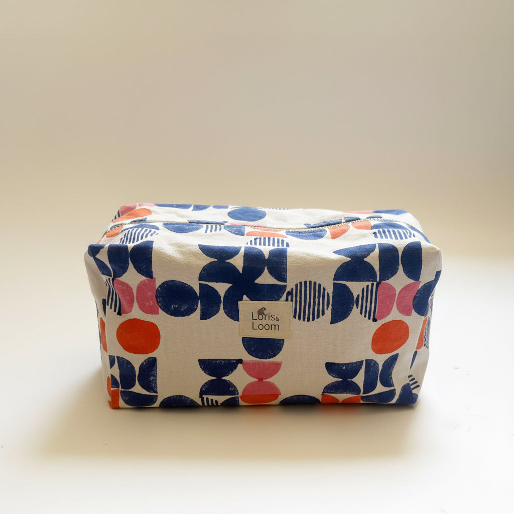 A rectangular zippered pouch with a white background and a colorful mid-century modern geometric pattern in indigo blue, coral pink, and orange. A small fabric label on the front reads 'Loris & Loom'.