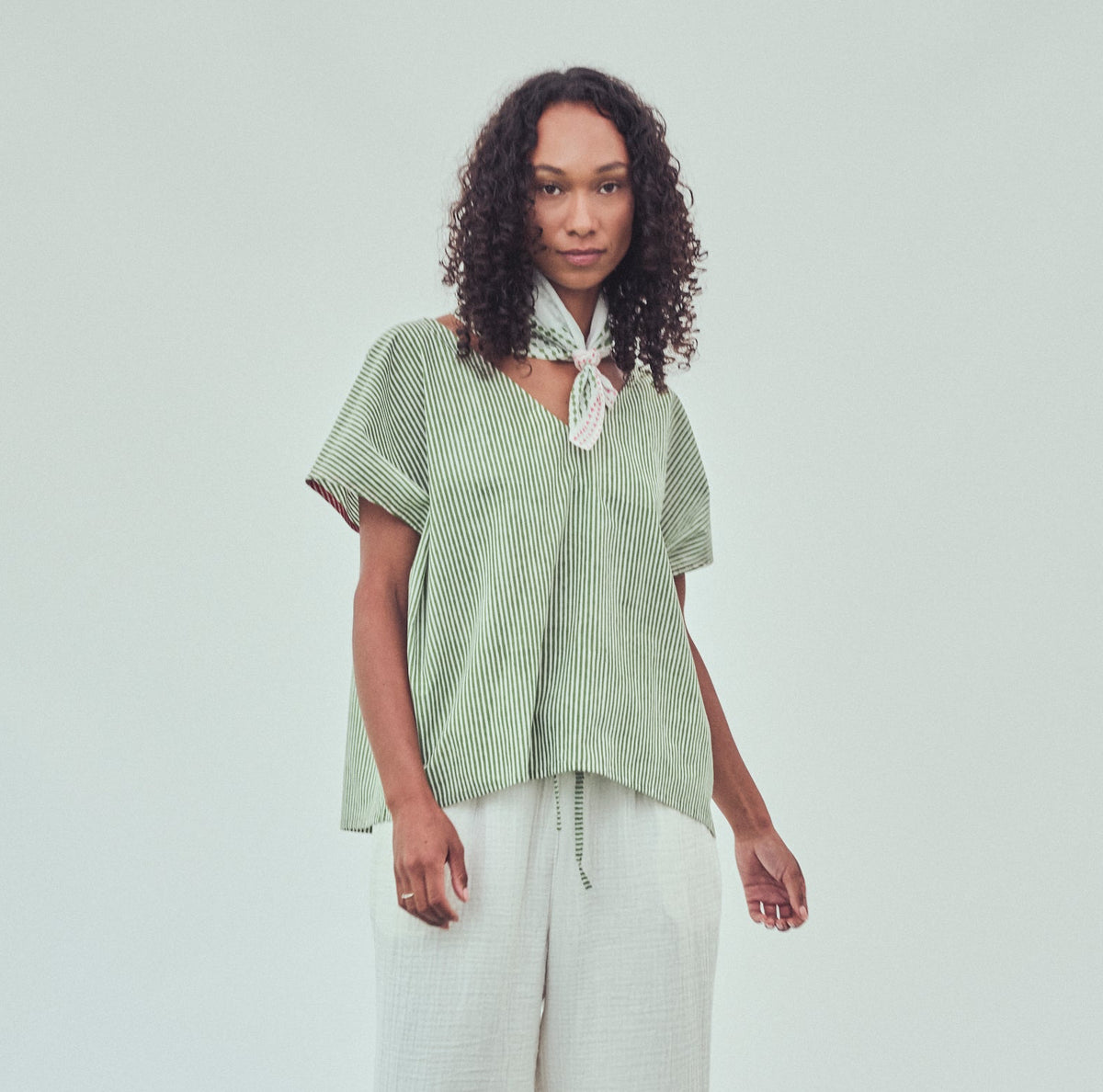 A model wearing a light sage green striped short-sleeve top with a tied neckerchief and  white wide-leg pants.