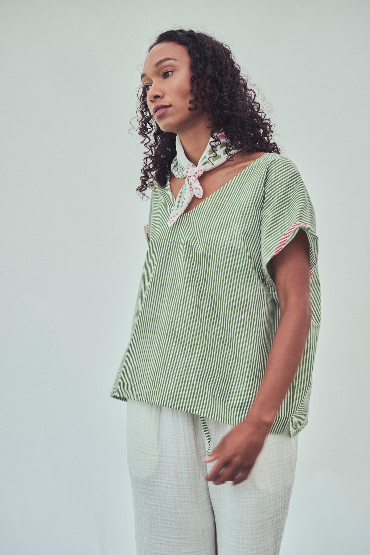 A woman wearing a light green and white vertically striped short-sleeve top with a white neck scarf, paired with white textured trousers, photographed against a plain light gray studio background.