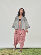 A model stands outdoors against a white studio backdrop on a strip of grass, wearing a black and white patterned open-front jacket, loose red striped pants, and sandals.