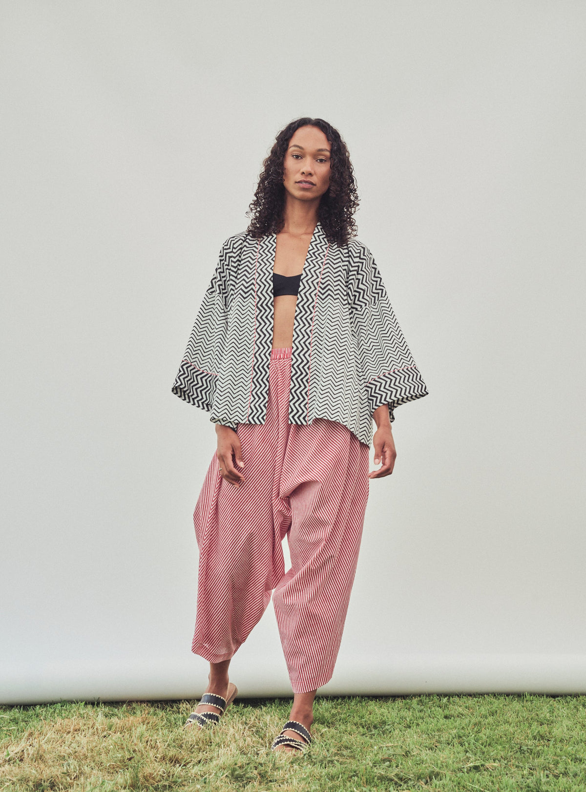 A model stands outdoors against a white studio backdrop on a strip of grass, wearing a black and white patterned open-front jacket, loose red striped pants, and sandals.