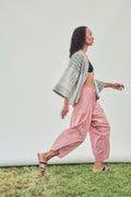 A woman walking in profile outdoors on grass, wearing a black-and-white patterned open-front jacket, a black crop top, red and white striped harem pants, and flat sandals against a plain light wall.