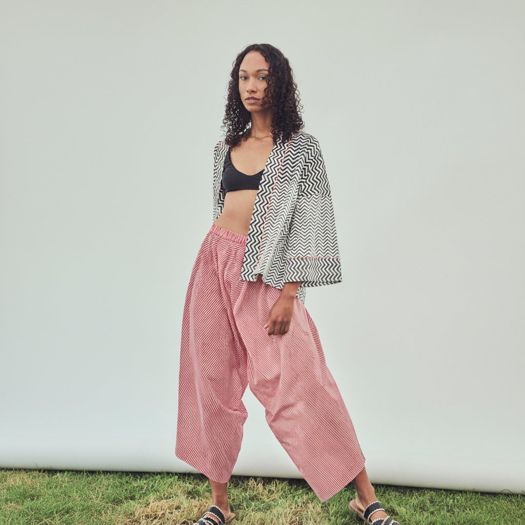 A model stands outdoors against a white backdrop wearing a black and white chevron-patterned open-front jacket, red and white striped harem  pants, a black bandeau top, and slide sandals.