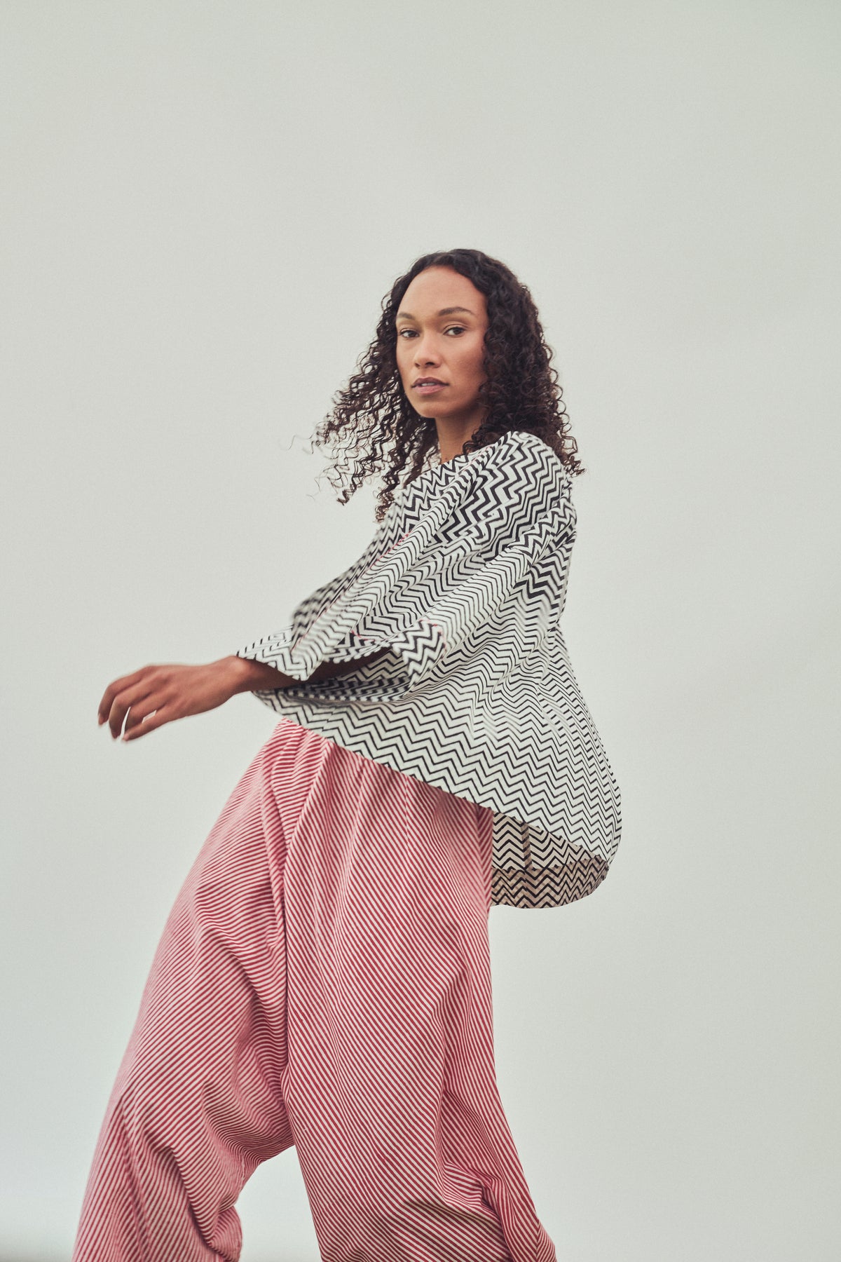 A model wearing a black and white chevron-patterned long-sleeve top and red-and-white striped harem pants, posed against a solid light grey studio background.