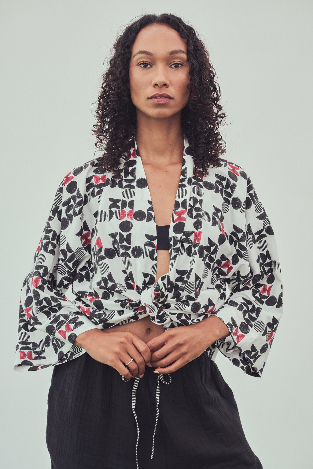 A woman wearing a kimono style jacket tied at the front with a black and burgundy abstract pattern. Tied at the waist over a black bandeau top and black drawstring pants, against a light gray studio 