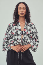 A woman wearing a kimono style jacket tied at the front with a black and burgundy abstract pattern. Tied at the waist over a black bandeau top and black drawstring pants, against a light gray studio 