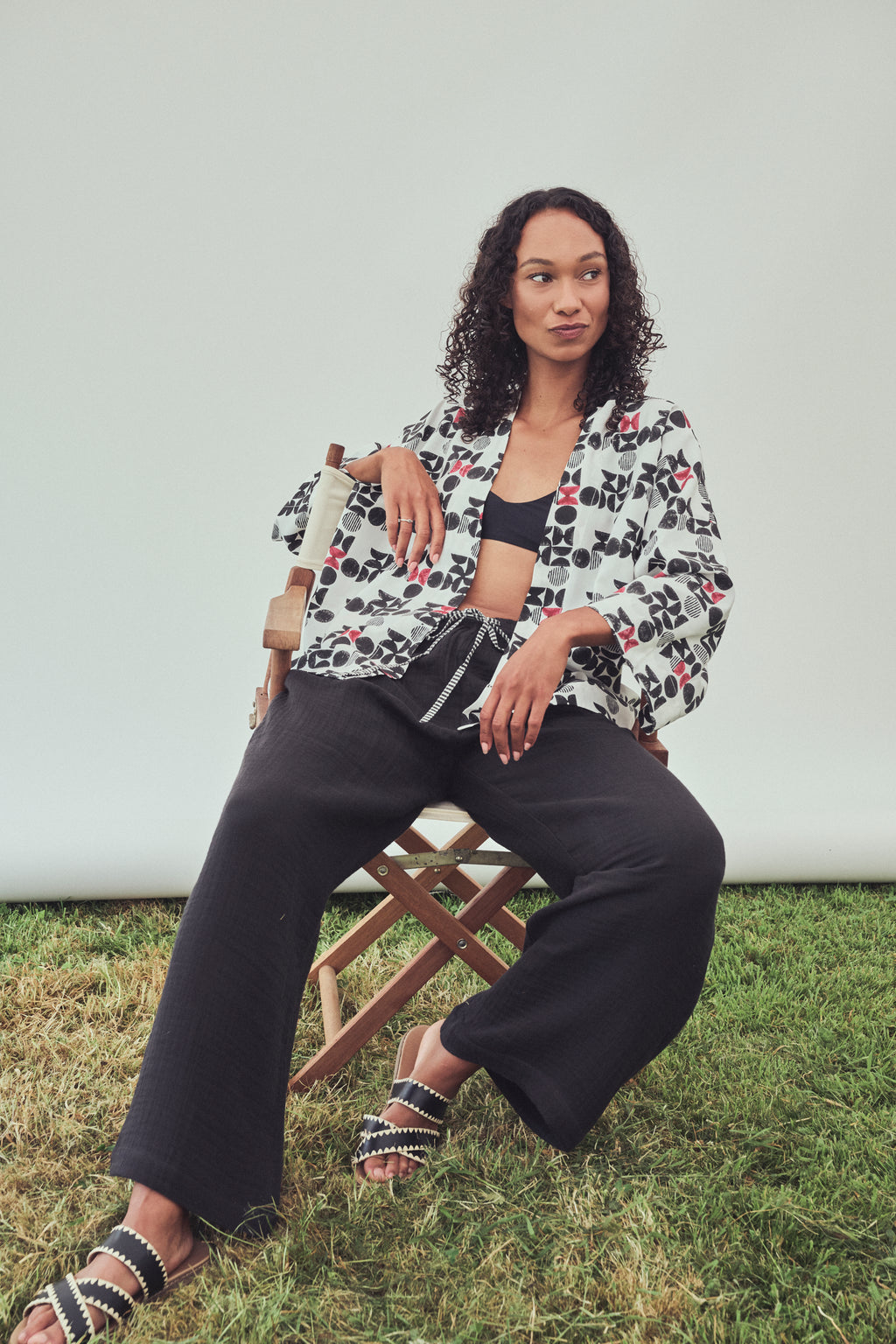 A woman sits on a wooden folding chair , wearing a black, white and red geo print kimono style jacket over a black bandeau top with black wide-leg pants, and black-and-white patterned slide sandals against a plain light gray backdrop.