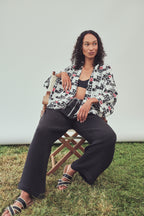 A woman sits on a wooden folding chair , wearing a black, white and red geo print kimono style jacket over a black bandeau top with black wide-leg pants, and black-and-white patterned slide sandals against a plain light gray backdrop.