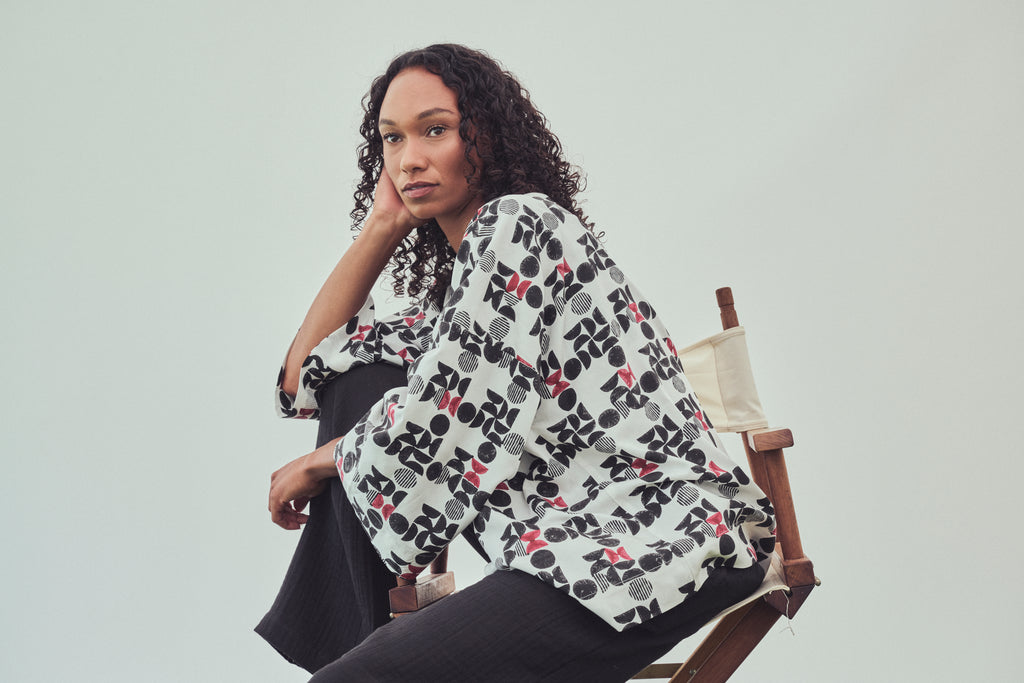 A woman with curly dark hair sits in a wooden chair against a light gray studio background, wearing a white kimono style jacket with a black and burgundy  geo pattern and black Trousers.