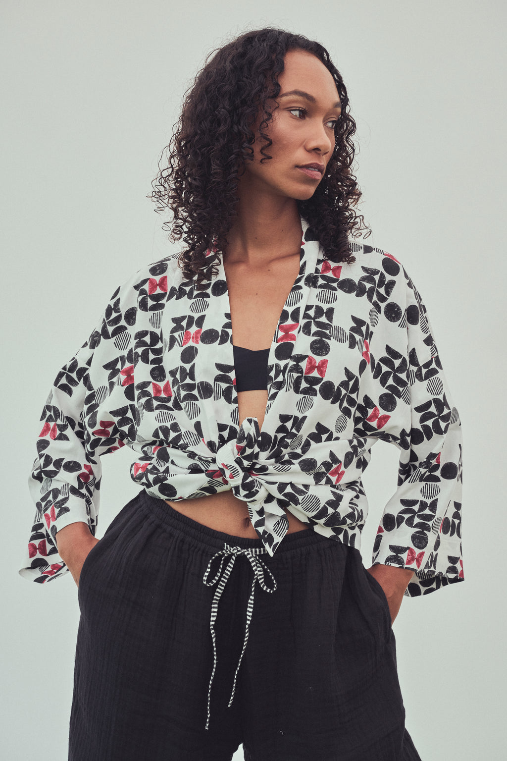 A woman wearing a kimono style jacket tied at the front with a black and burgundy abstract pattern. Tied at the waist over a black bandeau top and black drawstring pants, against a light gray studio background.
