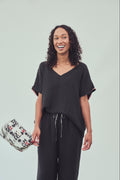 A woman modelling a black short-sleeve V-neck top and matching black drawstring pants against a light grey studio background, holding a patterned washbag.