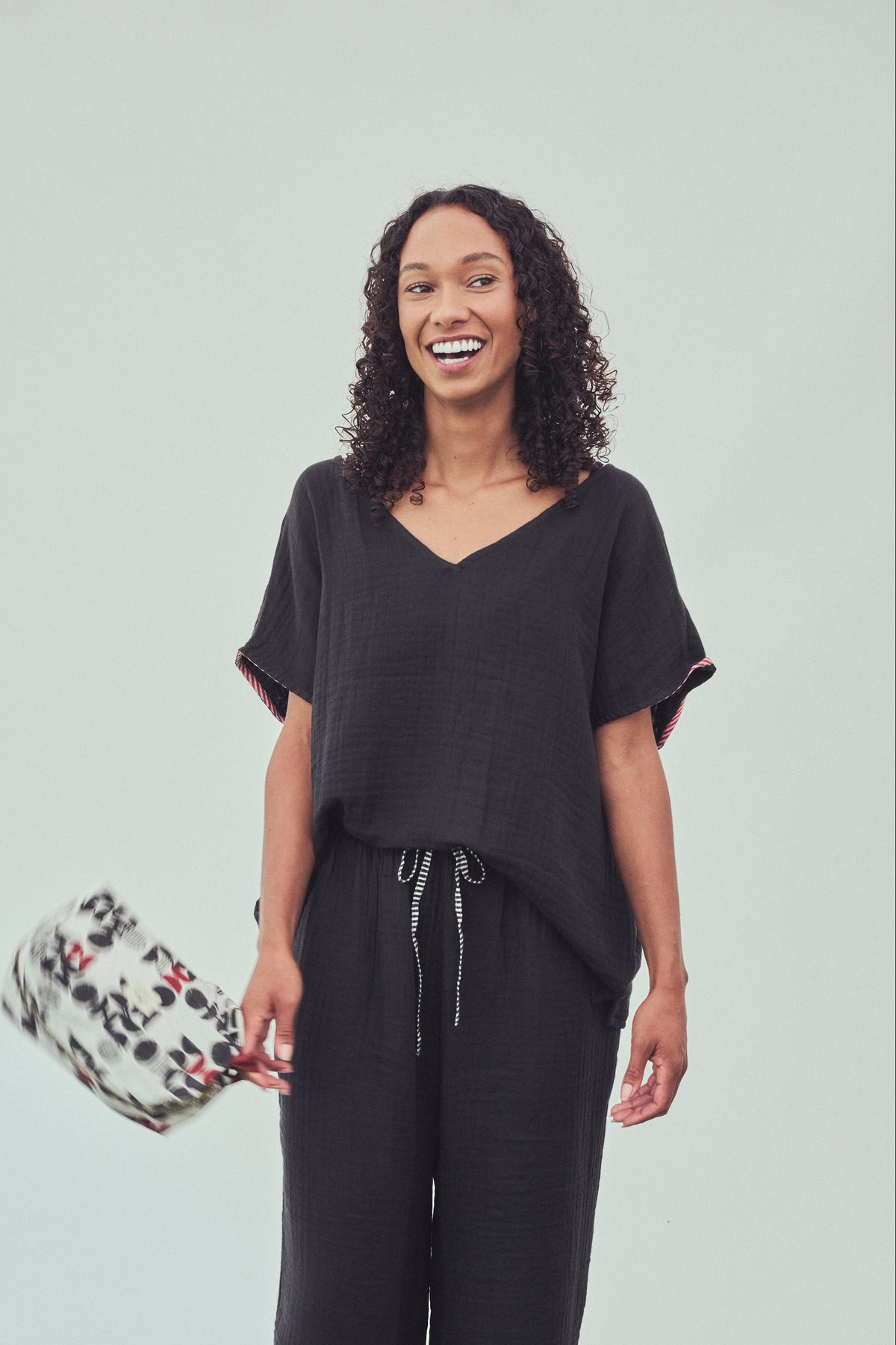 A woman modelling a black short-sleeve V-neck top and matching black drawstring pants against a light grey studio background, holding a patterned washbag.