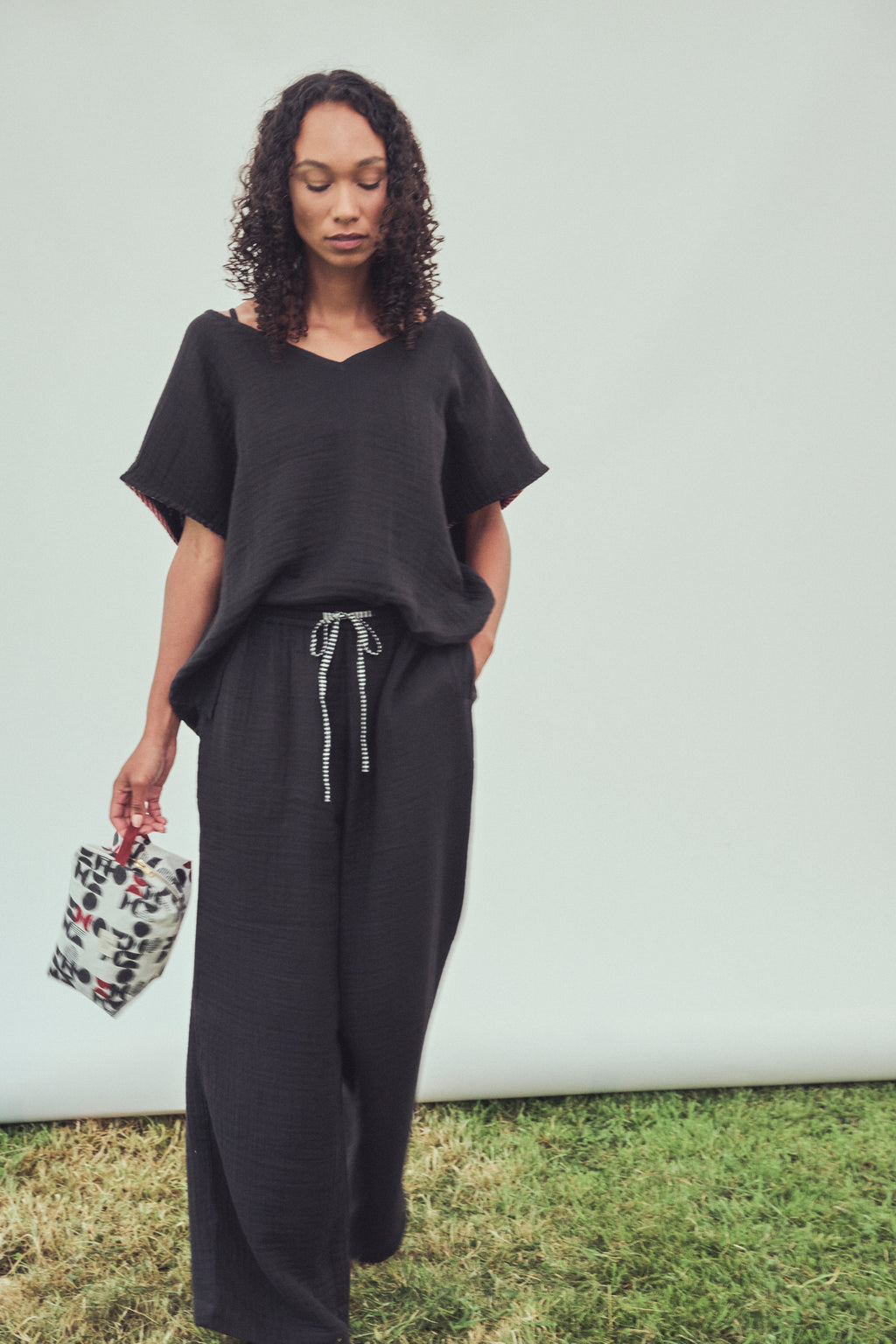 A woman stands outdoors against a white wall and grass, wearing a loose black short-sleeve top and matching black wide-leg pants with a drawstring waist, while holding a small patterned washbag.
