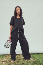 A woman stands outdoors against a light gray wall and grass, wearing a loose black short-sleeve top and matching black wide-leg drawstring pants, holding a patterned washbag.
