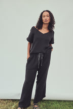 A woman modeling a black two-piece loungewear set consisting of a short-sleeve V-neck top and matching wide-leg drawstring pants, standing against a light gray backdrop.