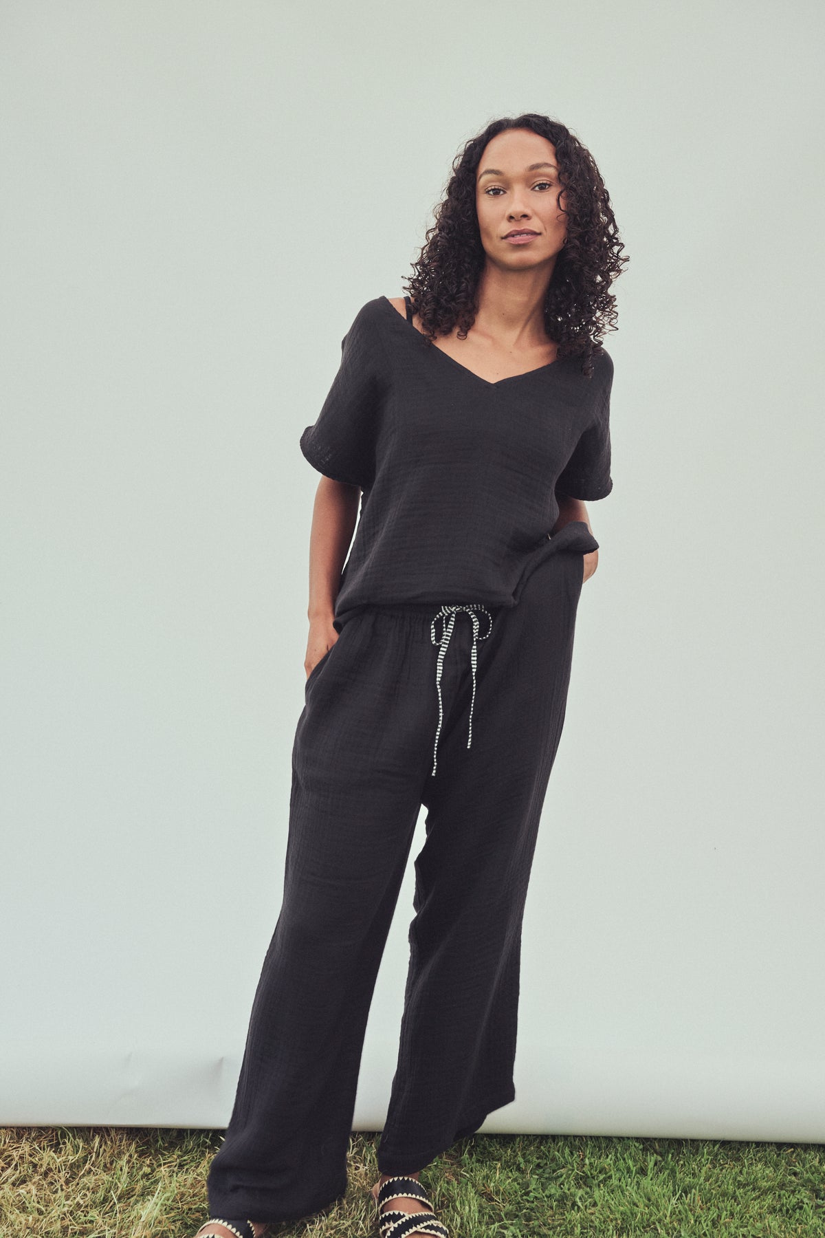 A woman modeling a black two-piece loungewear set consisting of a short-sleeve V-neck top and matching wide-leg drawstring pants, standing against a light gray backdrop.