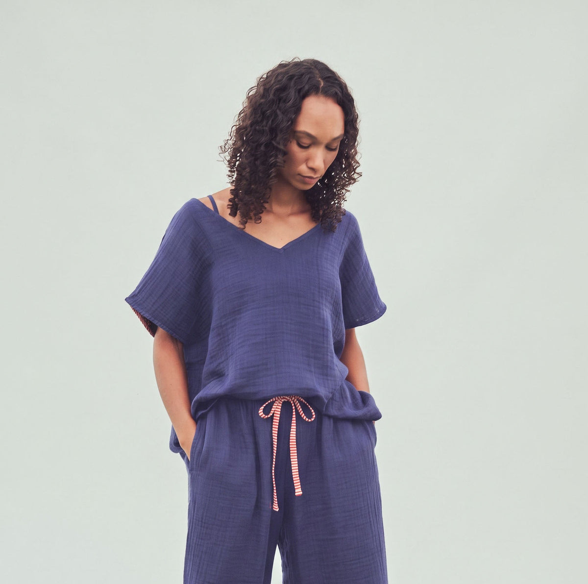 A woman wearing a matching two-piece lounge set in textured indigo blue cotton fabric, featuring a short-sleeve V-neck top and wide-leg pants with a red and white striped drawstring, standing outdoors on grass against a light gray backdrop.