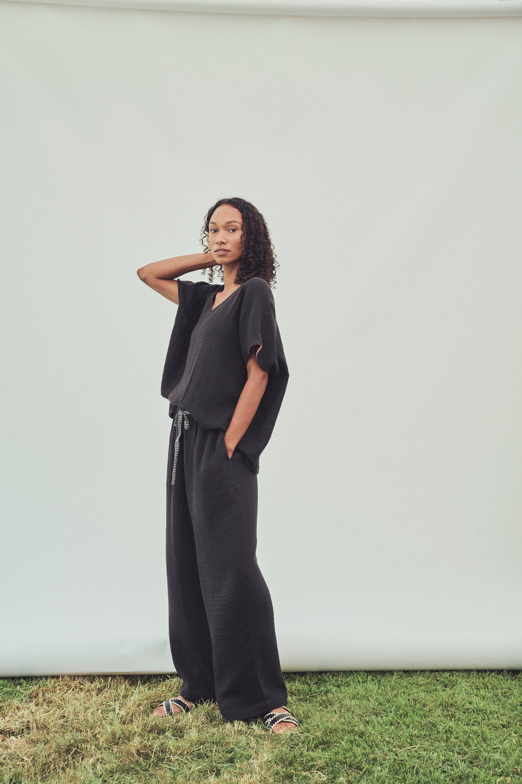 A woman modelling a black top with short sleeves and a drawstring waist trouser, standing on grass against a white studio backdrop.