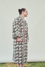 A man wearing a long-sleeved patterned robe with a geometric black and red print on a light background, s on grass against a plain light grey backdrop.