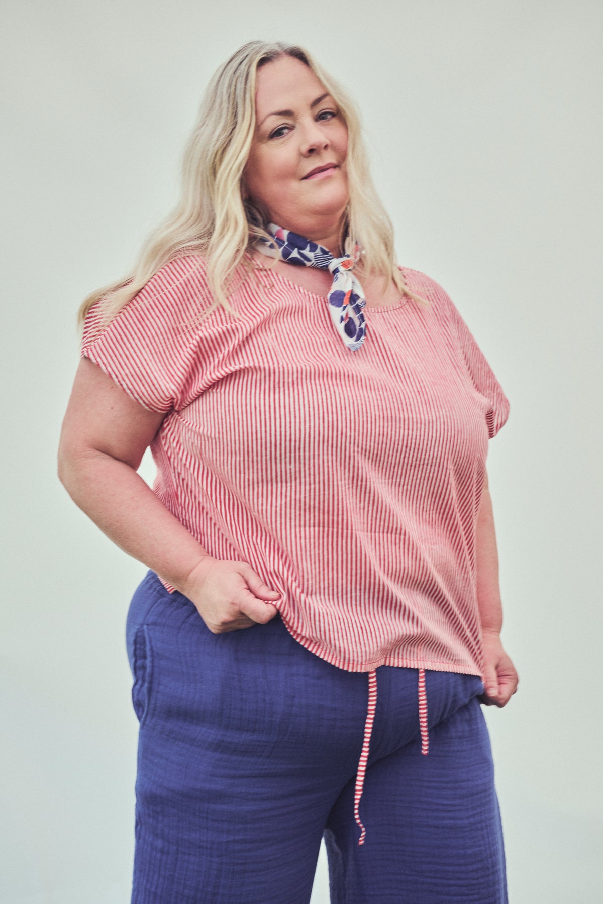 A woman modeling a short-sleeve red and white vertically striped top with a patterned neck scarf, paired with blue pants, against a plain light gray background.