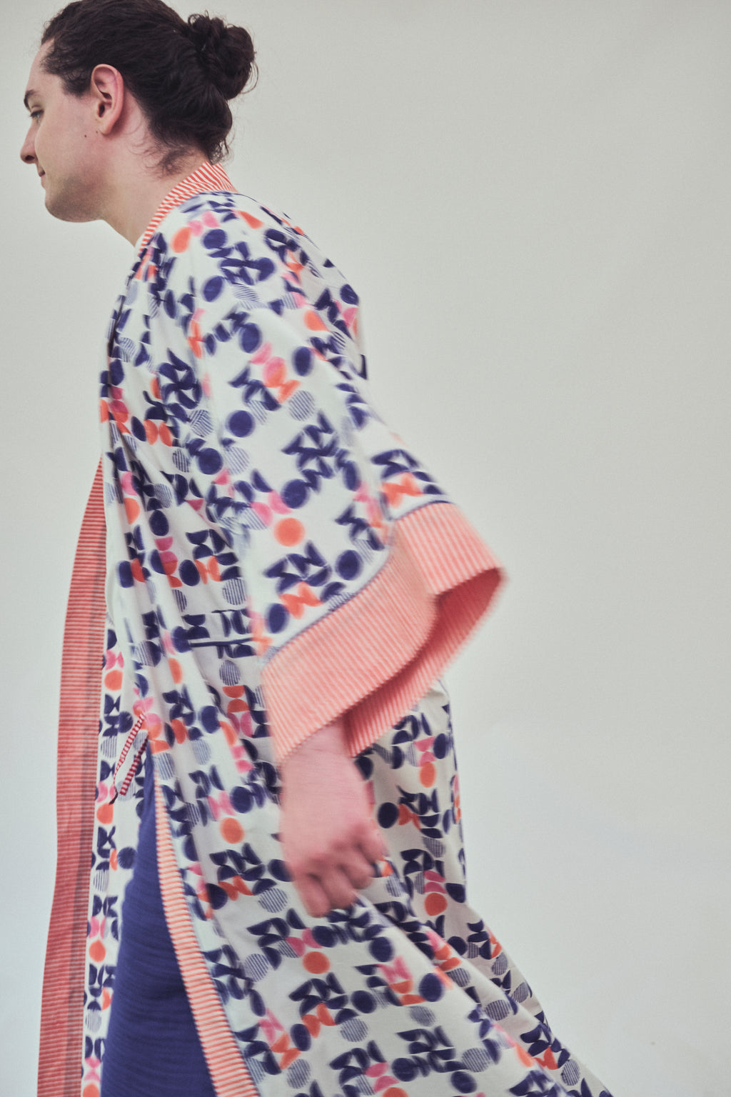A model shown from the side wearing a white kimono-style robe with a colorful abstract pattern in indigo, orange, and white, featuring orange striped trim.