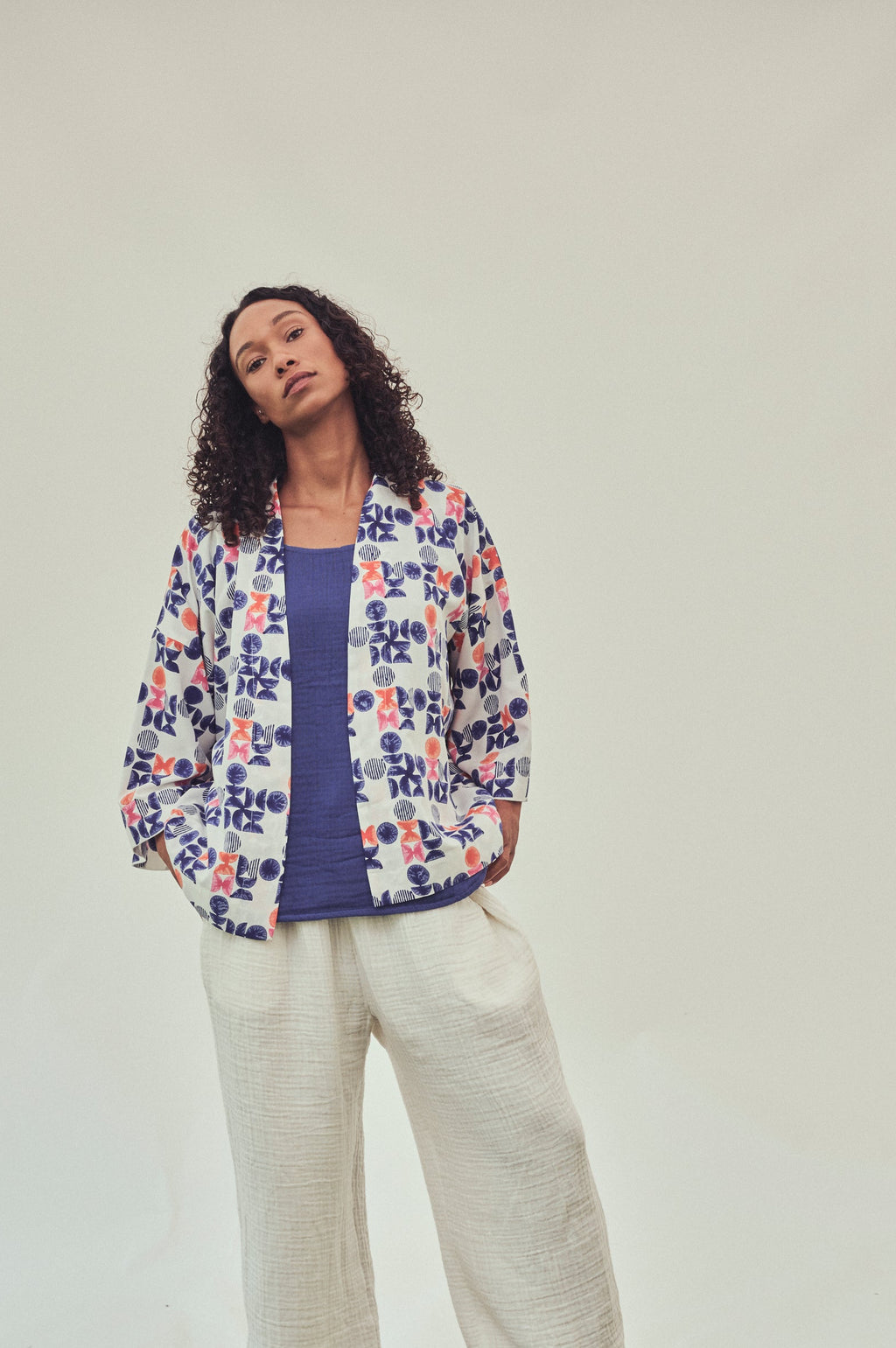 A woman stands outdoors against a white backdrop, wearing a printed geo  jacket over an indigo top and ivory wide-leg pants.