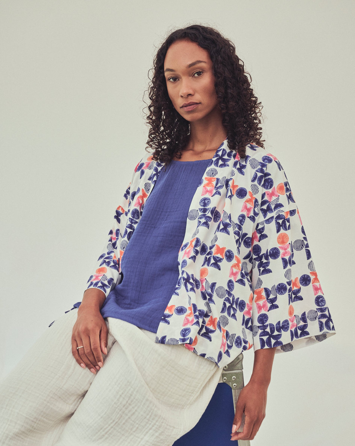 A woman models a printed three-quarter sleeve jacket with a colourful geo print in indigo, orange, and pink, on a white ground. Worn over a solid indigo top and ivory-coloured textured trousers.