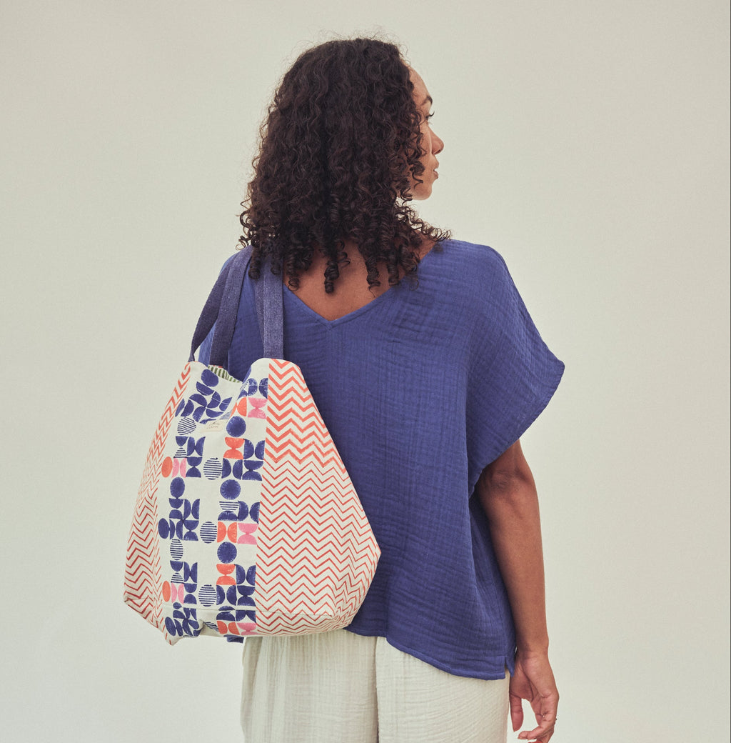 A woman shown from the back wearing an indigo top and ivory pants, carrying a large patterned tote bag on her shoulder. The fabric bags have an ivory base with blue, pink and orange  geometric print, set against a plain light background.