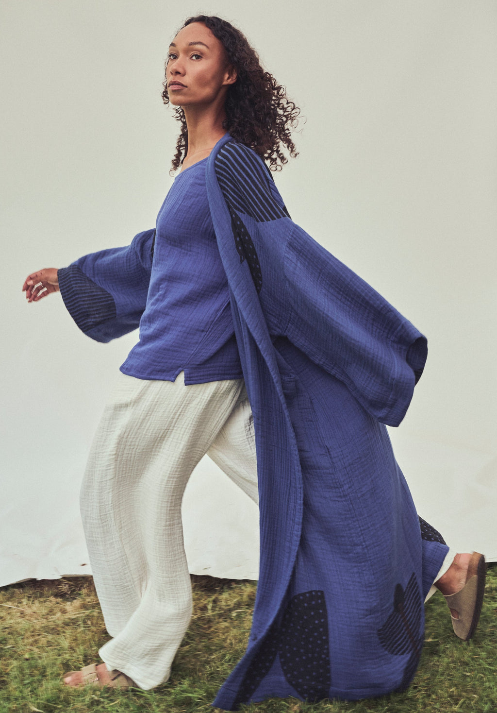 A woman wearing a short Indigo top and long loose, oversized indigo robe and white textured wide-leg pants, standing outdoors on grass in front of a white fabric backdrop.