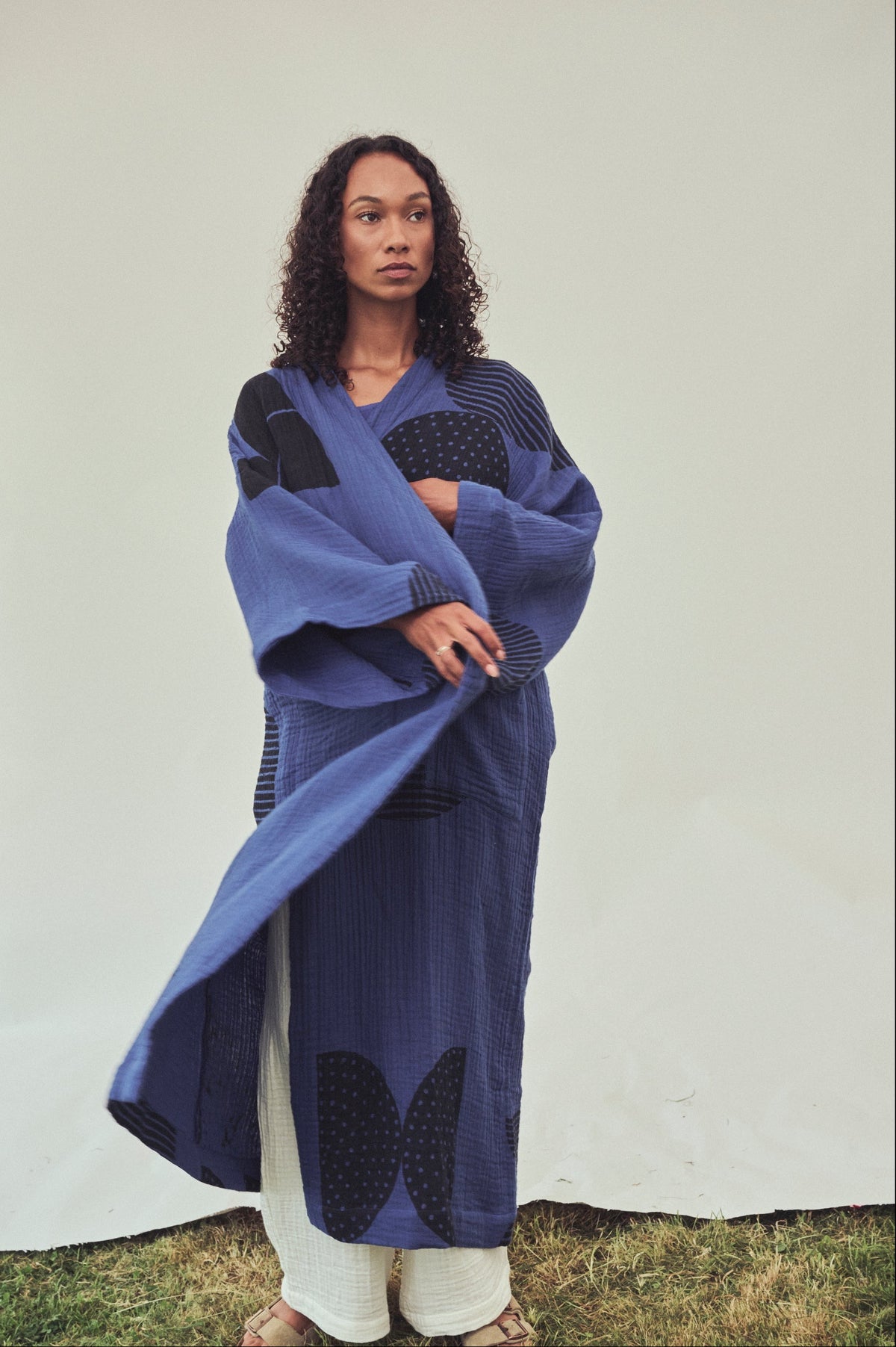 A woman stands outdoors wearing a long blue kimono-style robe with wide sleeves, layered over white pants. The robe features a black abstract print 