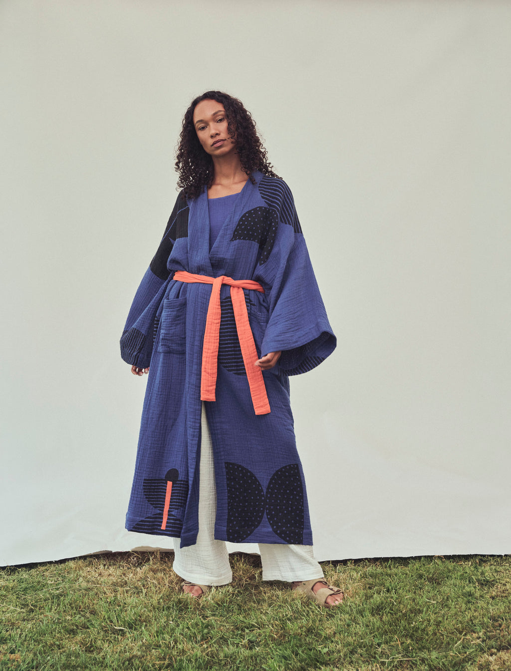 A model stands outdoors on grass wearing a loose, long-sleeved kimono-style robe in blue and black with an orange belt, paired with white pants and sandals.