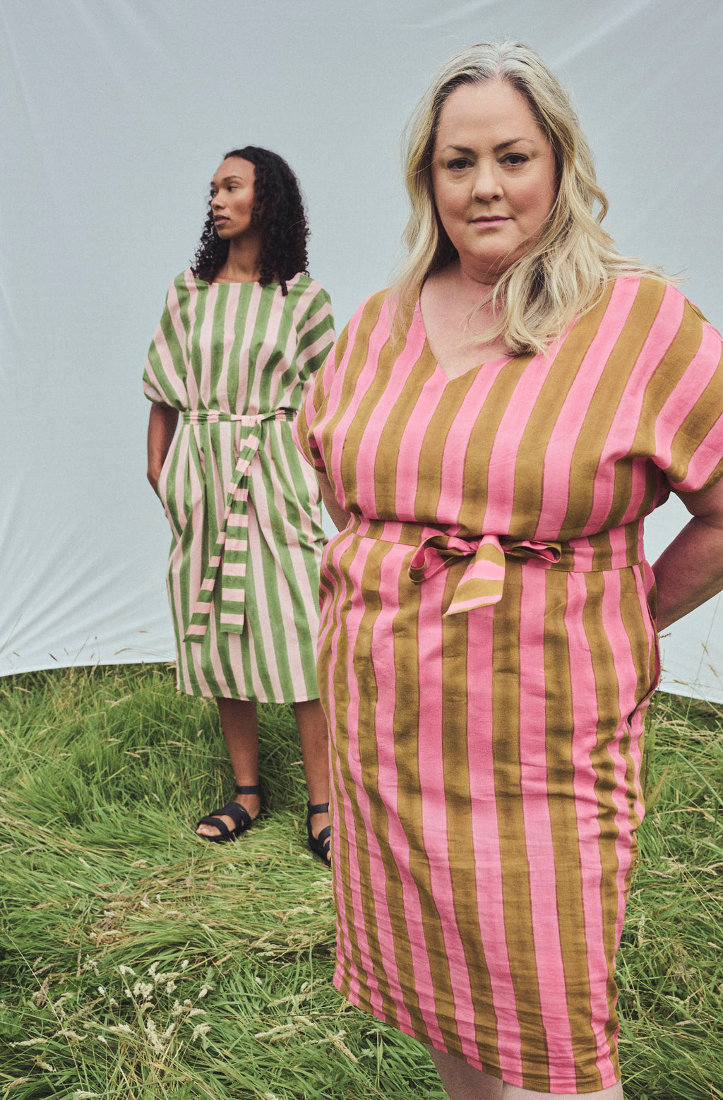 Two women modeling striped midi dresses outdoors on grass in front of a white tent. The woman in the foreground wears a pink and ochre vertically striped dress with a tie waist, while the woman behind wears a green and pale pink striped dress.