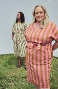 Two women modeling striped midi dresses outdoors on grass in front of a white tent. The woman in the foreground wears a pink and ochre vertically striped dress with a tie waist, while the woman behind wears a green and pale pink striped dress.