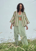 A model stands outdoors in front of a large white fabric backdrop, wearing a light green and pink striped open-front jacket with wide sleeves and striped green and white harem pants.