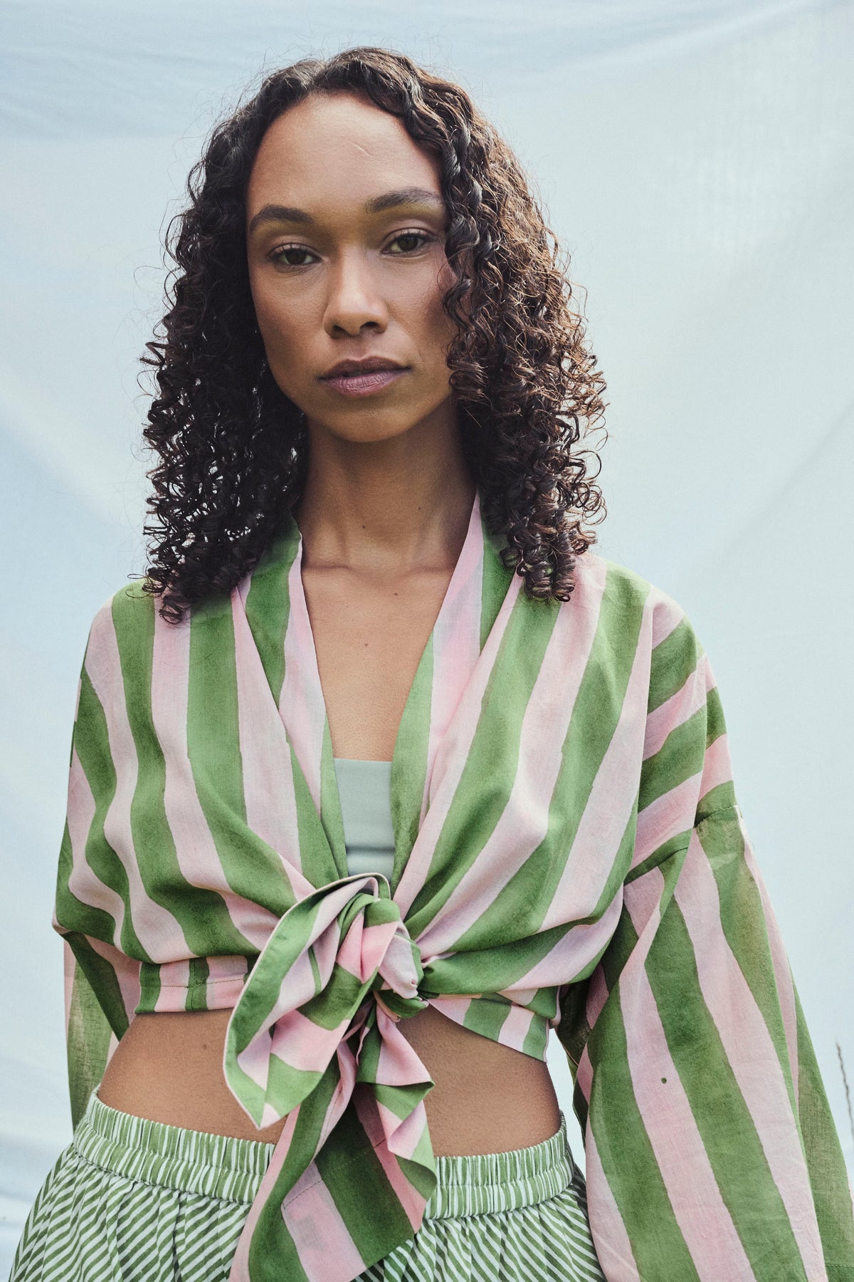 A model with curly dark hair wearing a light green and pink striped top tied at the waist, paired with striped bottoms, against a soft light grey background.