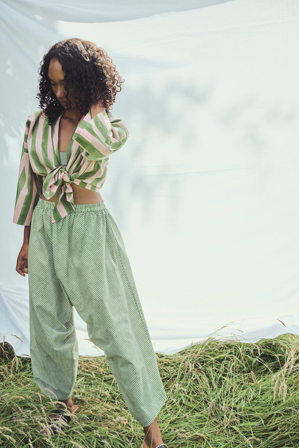 A model wearing a light green and pink striped top tied at the waist and  green and white striped harem balloon pants, standing outdoors on  grass against a white fabric backdrop.