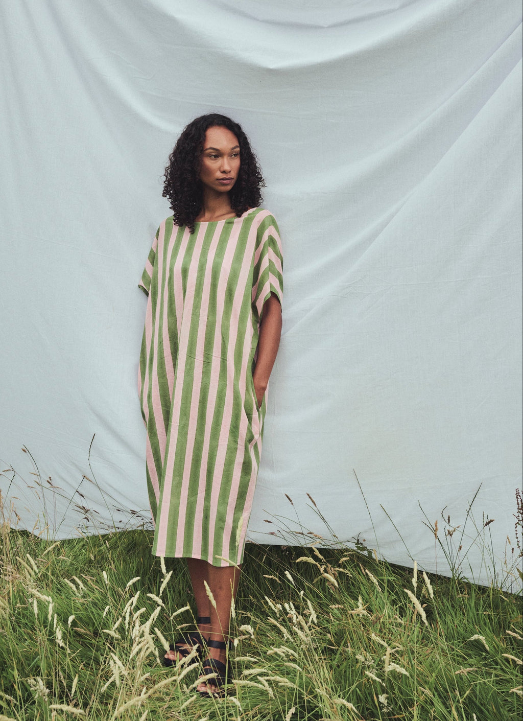 A woman stands outdoors in a field in front of a large white fabric backdrop, wearing a loose, knee-length dress with vertical sage green and pale pink stripes.