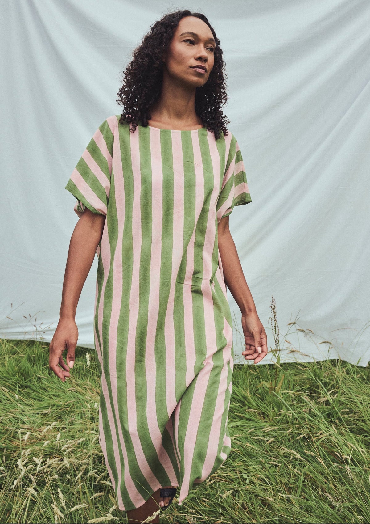 A woman wearing a loose, short-sleeved midi dress with vertical sage green and light pink stripes, standing in a grassy field in front of a white fabric backdrop.