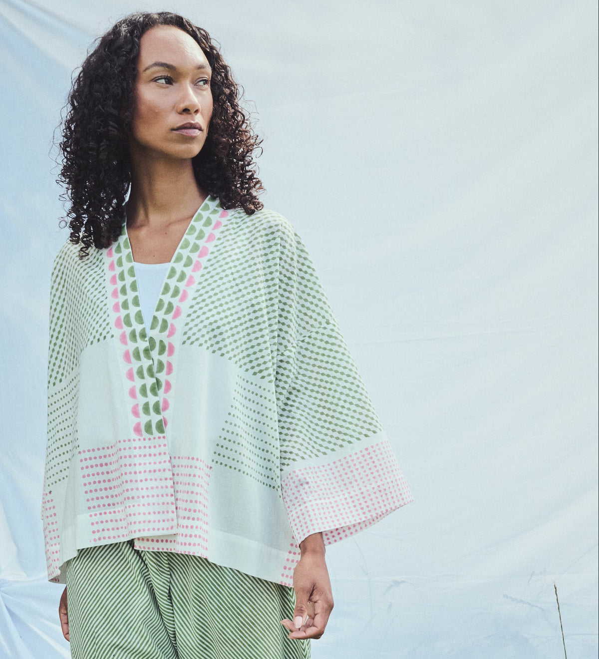 A woman wearing a light green, pink and white patterned kaftan-style top, paired with green striped trousers, standing against a light grey fabric backdrop.