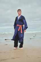 A man walking on a sandy beach wearing a blue and black printed kimono-style robe with an orange belt and black pants, under an overcast sky.