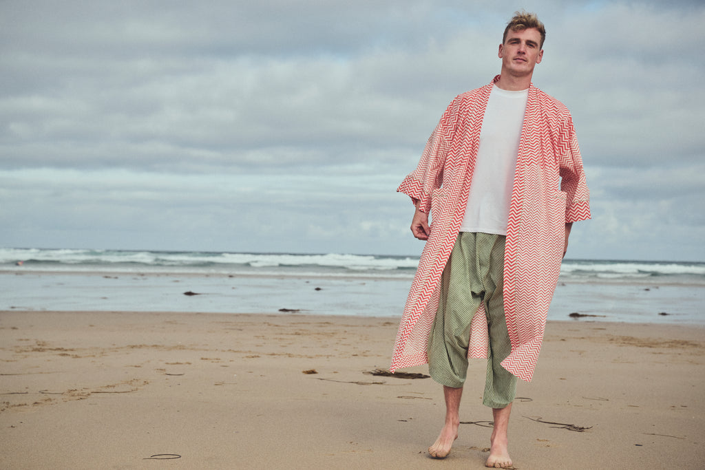 A man standing on a sandy beach wearing a long-sleeved, open-front orange and white zigzag robe over a white t-shirt and  green and white striped harem cropped pants, with the ocean and cloudy sky in the background.
