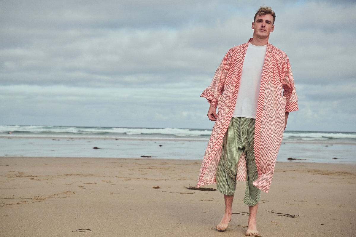 A man standing on a sandy beach wearing a long-sleeved, open-front orange and white zigzag robe over a white t-shirt and  green and white striped harem cropped pants, with the ocean and cloudy sky in the background.