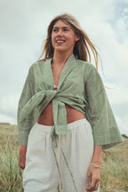 A woman standing outdoors in  grassy sand dunes wearing a light sage green and white striped wrap-style top tied at the waist, paired with white drawstring pants.