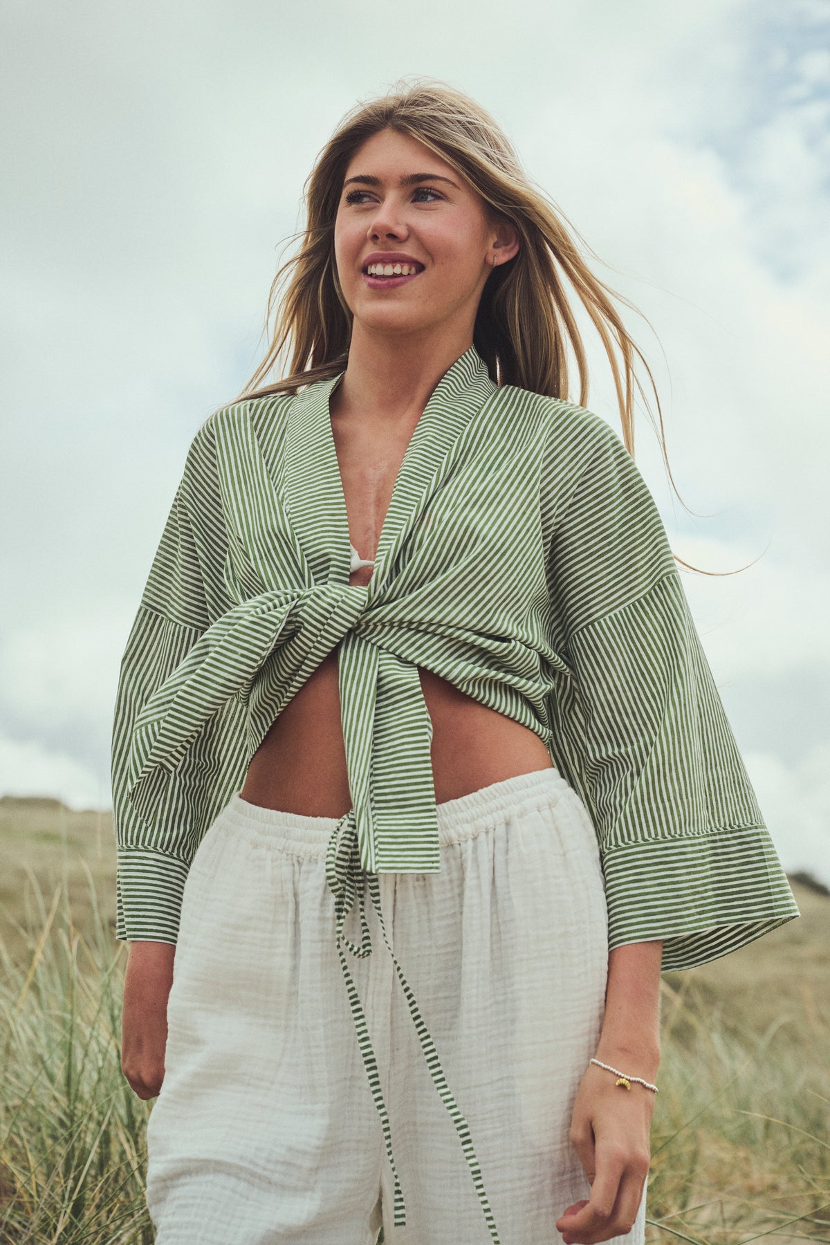 A woman standing outdoors in  grassy sand dunes wearing a light sage green and white striped wrap-style top tied at the waist, paired with white drawstring pants.