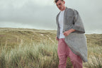 A man standing on grassy dunes under an overcast sky, wearing a white t-shirt, red striped harem pants, and a black and white chevron-patterned robe.