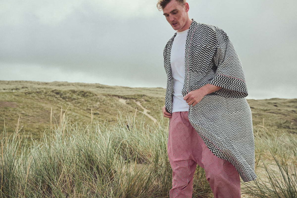 A man standing on grassy dunes under an overcast sky, wearing a white t-shirt, red striped harem pants, and a black and white chevron-patterned robe.