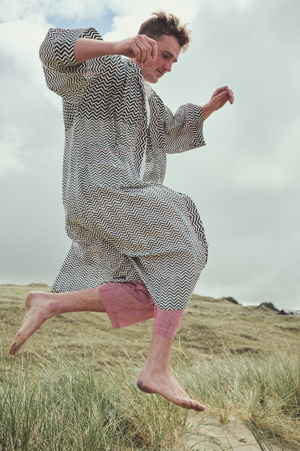 A man jumps barefoot on grassy dunes wearing a loose, long-sleeve kaftan-style robe with a black and white zig-zag pattern, paired with red striped pants.