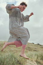 A man jumps barefoot on grassy dunes wearing a loose, long-sleeve kaftan-style robe with a black and white zig-zag pattern, paired with red striped pants.