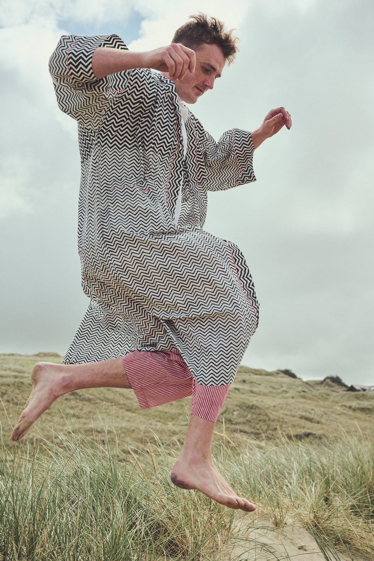 A man jumps barefoot on grassy dunes wearing a loose, long-sleeve kaftan-style robe with a black and white zig-zag pattern, paired with red striped pants.
