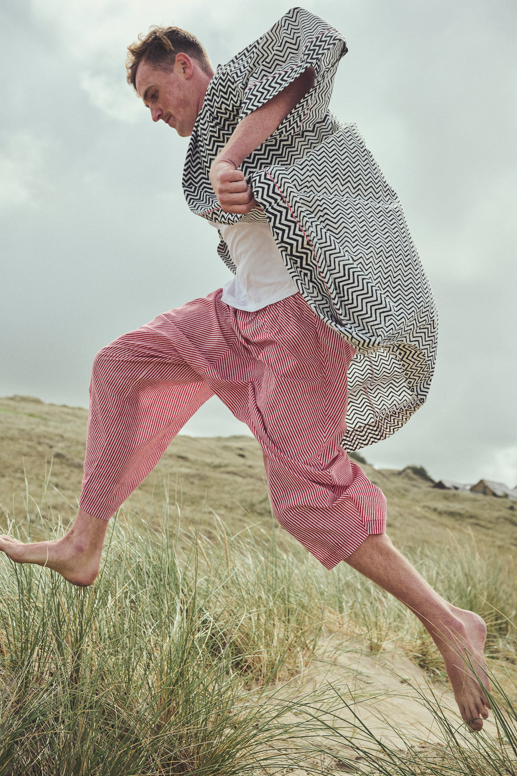 A man running barefoot through grassy dunes, wearing a black and white zigzag-patterned robe, a white Tshirt, and red striped harem pants.